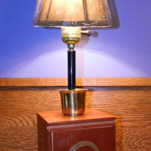 This lamp is an upright vertical piece created from a wooden Last Call cigar Box. It has a metal "shot glass" at the base of the stem and sports a small tan cloth shade on top.