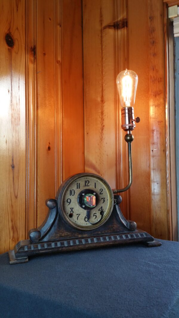 A mantle clock with Nixie tube in the middle of the clock face and a upturned arm coming out of the side topped by copper socket and an Edison-style light bulb.