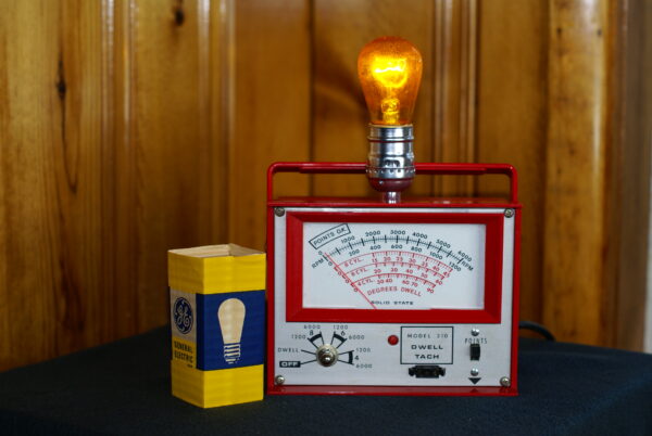 Made for the motorhead in your life. A red automobile diagnostic meter made into a lamp with a yellow light bulb on top with vintage light bulb packaging shown.
