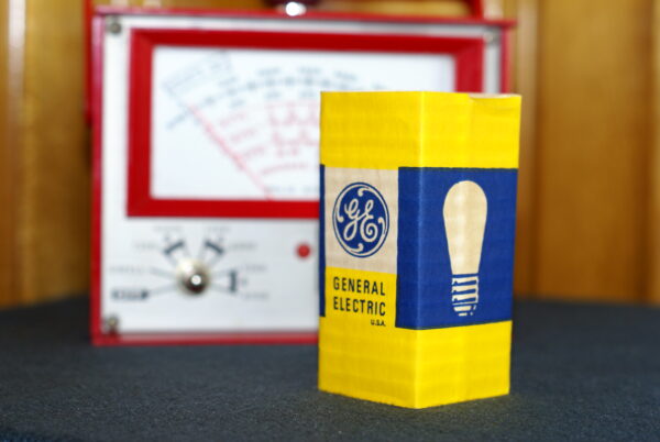 Made for the motorhead in your life. The back of of a red automobile diagnostic meter made into a lamp with a yellow light bulb on top with vintage light bulb packaging shown in a close-up..