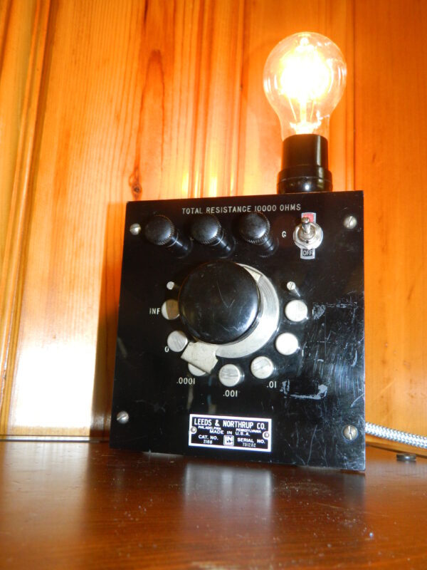 A heavy bakelite piece of vintage test equipment with a large black center dial, and several heavy metal silver contacter plates that the large center knob revolve around. A toggle switch to turn the light on and off, and a cloth wrapped vintage looking power cord.