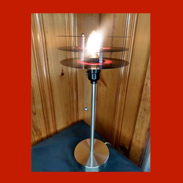 A silver pedestal style lamp topped by 3 red labeled 45 records that form the shade around a LED light.