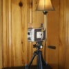 A brown and silver Kodak 8 mm Movie camera with a light fixture mounted to it with a small shade over the bulb. The camera is set upon a tripod.