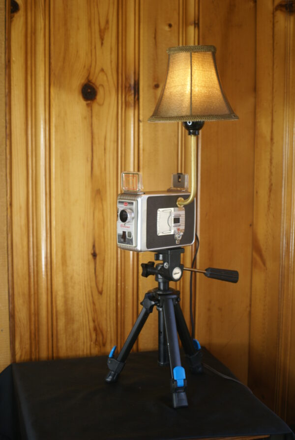 A brown and silver Kodak 8 mm Movie camera with a light fixture mounted to it with a small shade over the bulb. The camera is set upon a tripod.
