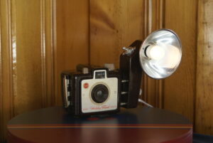 A small vintage Kodak camera with a flash unit attached to the side. The flash unit has been rewired with a toggle switch and light socket and a small white LED buld is not mounted where the flash bulb used to be. Small table top light.