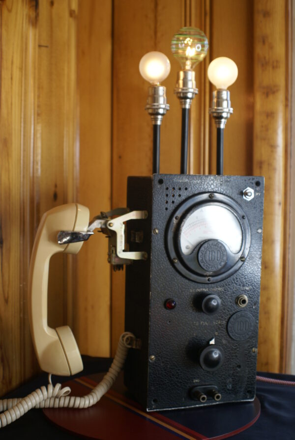a black in color 1930s voltmeter with a vintage telephone receiver hanging upon the left side. The meter has a broad face with a prominent gauge on the front reading "Volts" and various numbers on the face. There is a toggle switch on the front of the unit and three tall black stalks emerging from the top, which have light sockets upon each one..