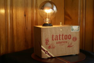 A natural wood cigar box lamp with a USB port installed on the back side of the lamp. A small gold rotator switch and a silver topped globe LED light bulb.