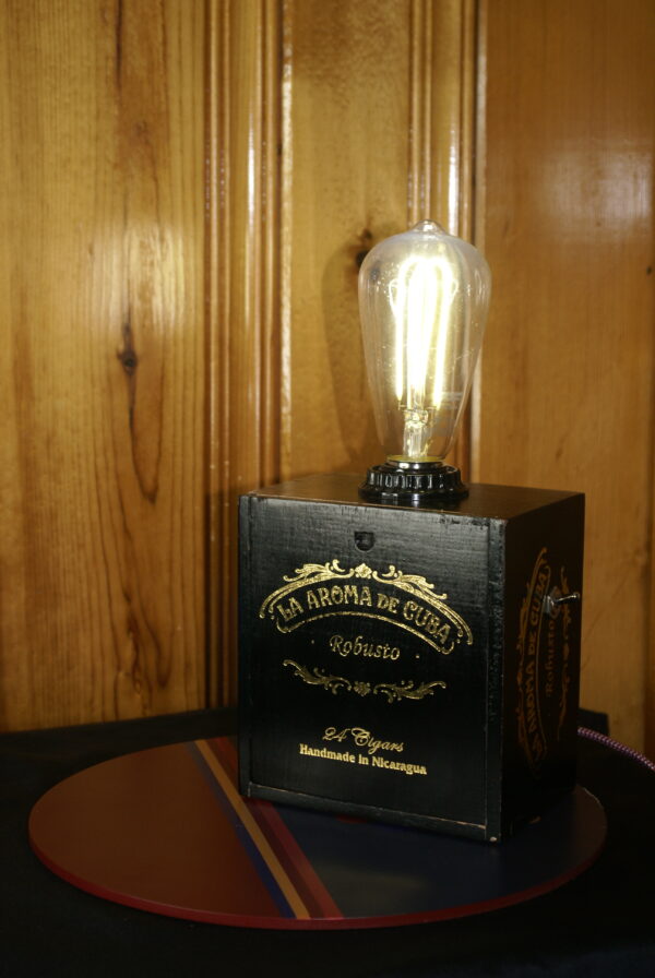 A black and gold colored cigar box lamp with gold script which reads La Aroma De Cuba, There is a vintage style LED Edison style light bulb and a cloth covered vintage style power cord.