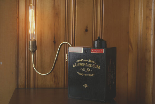 A black cigar box with gold lettering in script that reads La Aroma De Cuba. The box has a sweeping a curved arm that comes off to the left side upon there is a brass light socket with a long tubular shaped edison style light bulb. There is a switch on the top of the box that is round and vintage in appearance.